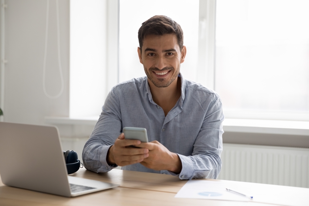 Young man updating his social media on his phone with an open laptop computer beside him.